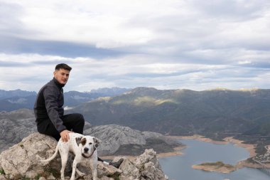 Dağın tepesinde karanlık dağ kıyafetleri giymiş genç dağcı köpeğinin yanındaki bir kayanın üzerinde dinleniyor. Arka planda bir göl ve komşu sıradağları görüyoruz.. .