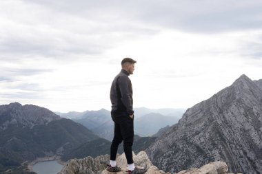 Karanlık dağ kıyafetleri giymiş genç dağcı dağın tepesindeki ufka doğru bakıyor. Arka plan manzarası nefes kesici ve dik dağlar görülebilir. Yürüyüş ve tırmanış için duvar kağıtları.