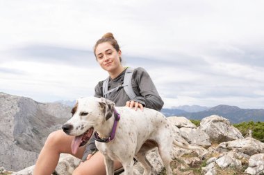 Dağ kıyafetleri ve gri bir sırt çantası giymiş gülümseyen beyaz bir kız. Bu macerada köpeği ona eşlik ediyor. Arka planda dağ sırasını görebilirsiniz. Yürüyüş ve tırmanış için duvar kağıtları.