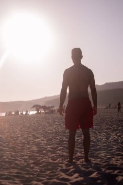 Koyu renk saçlı ve tenli genç bir adam plajda bir tatil gününde güneş ışığını gözlemler..