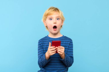 Akıllı telefonu tutan küçük bir çocuk, ağzı açık kalmış, şok olmuş. Çocuk ve telefonu, pastel mavi arka planda izole edilmiş. Çocuklar ve teknoloji, tehlikeler, ekran süresi ve ebeveyn denetim uygulamaları. 