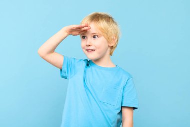 Hoş bir anaokulu öğrencisi, eli alnında ve gülümsüyor. Çocuk geleceğe bakıyor, stüdyo resmi mavi arka planda..