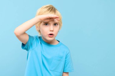 Sevimli anaokulu öğrencisi, ilginç bir şekilde eli alnında uzaklara bakıyor. Çocuk geleceğe bakıyor, stüdyo resmi mavi arka planda..