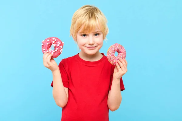 Cute little boy with blond hair and freckles having fun with glazed donuts. Children and sugary junk food concept. Boy holding colorful donuts, eating junk unhealthy food full of sugar, isolated on pastel blue studio background.