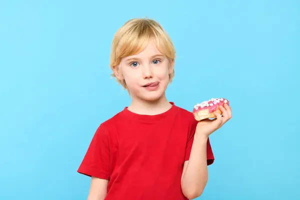 Cute little boy with blond hair and freckles having fun with glazed donuts, sticking tongue out. Children and sugary junk food concept. Boy holding colorful donuts, eating junk unhealthy food full of sugar, isolated on pastel blue studio background.