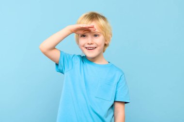 Hoş bir anaokulu öğrencisi, eli alnında ve gülümsüyor. Çocuk geleceğe bakıyor, stüdyo resmi mavi arka planda..