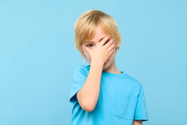 Duygusal genç çocuk, elleri pastel mavisi arka planda soyutlanmış olarak yüzünü kapatıyor. Çocukluk çağında zihinsel refah ve güvenlik.