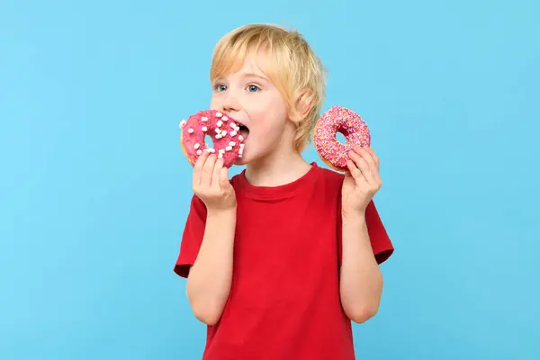 かわいい男の子 ブロンドの髪とフリックル 艶をかけられたドーナツを食べる 子供と砂糖のジャンクフードコンセプト カラフルなドーナツを持っている少年 パステルブルーのスタジオの背景に隔離された砂糖でいっぱいのジャンク不健康な食べ物を食べる ロイヤリティフリーのストック画像