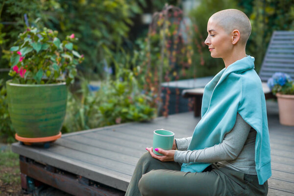 Молодая пациентка с раком сидит в саду с кофе, глаза закрыты. Психическое здоровье, благополучие, баланс жизни, медитация во время болезни.