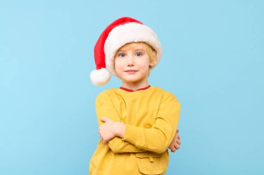 Noel Baba şapkası takan, kameraya bakarken gülümseyen beyaz bir çocuk. Stüdyo portresi mavi arkaplanda izole edildi.