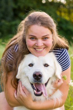 Beautiful young woman hugging her cute large white Great Pyrenees dog laying in tall grass meadow, enjoying time together, smiling and looking at camera. clipart