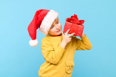 Kırmızı Noel Baba şapkası takan yakışıklı çocuk Noel hediyesini tutuyor. Pastel mavisi arka planda izole edilmiş bir stüdyo resmi. Tatil arkaplanı.
