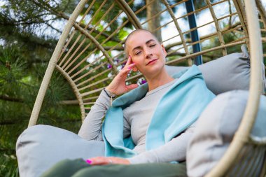 Young female cancer patient sitting on garden patio, rocking in a hanging chair. Health insurance, medical expenses, loss of income safety plan.  clipart