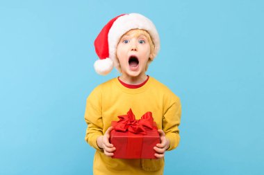Kırmızı Noel Baba şapkası takan yakışıklı çocuk Noel hediyesini tutuyor. Pastel mavisi arka planda izole edilmiş bir stüdyo resmi. Tatil arkaplanı.