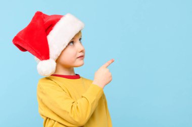 Noel Baba şapkası takan sevimli çocuk pastel mavi arka planda parmağıyla bir tarafı işaret ediyor. Promosyon reklamı için kopya alanı gösteren anaokulu öğrencisi. 