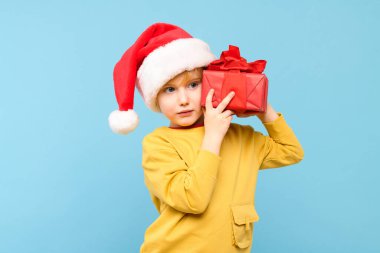 Kırmızı Noel Baba şapkası takan yakışıklı çocuk Noel hediyesini tutuyor. Pastel mavisi arka planda izole edilmiş bir stüdyo resmi. Tatil arkaplanı.