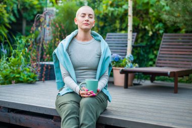Bahçe avlusunda oturan genç bayan kanser hastası, gözleri kapalı kahve içiyor. Akıl sağlığı, refah, yaşam dengesi, hastalık sırasında meditasyon..