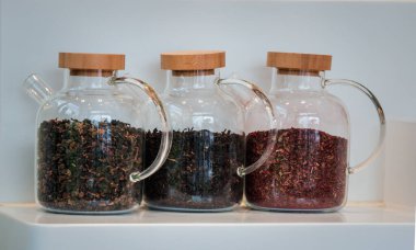 Beyaz bir rafta, içinde üç çeşit kurutulmuş çay yaprağı bulunan ahşap mantar kapaklı üç cam kavanoz var..