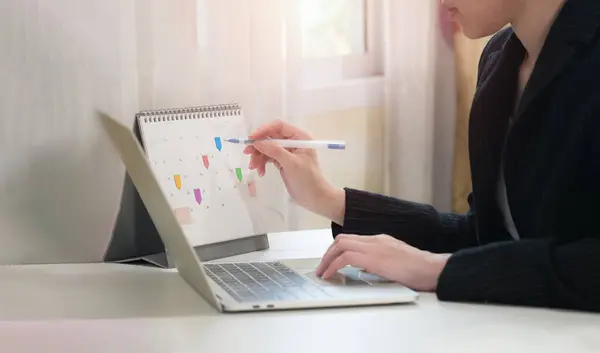 Event planner calendar, agenda, timetable for 2024. Businesswoman using phone to check planner and making notes on desk calendar at her desk in the office. Work planning, event calendar.