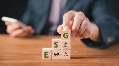 ESG, sürdürülebilir bir işletmenin çevresel, sosyal ve idari yaratımı anlamına geliyor. Zorluk, bir Esg simgesi ve ilginç bir arkaplanda diğer simgeleri gösterirken tahta küpleri bir elde tutmaktır.