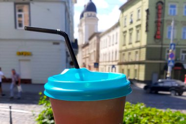 Üzerinde parlak mavi bir kapak ve siyah bir pipet bulunan kahverengi bir kahve bardağı ön planda belirgin bir şekilde sergilenirken, hareketli bir şehir arka planda sessizce sergilenir. Sahne yazın sıcak havasını yakalar, yoldan geçenleri eğlenmeye davet eder.