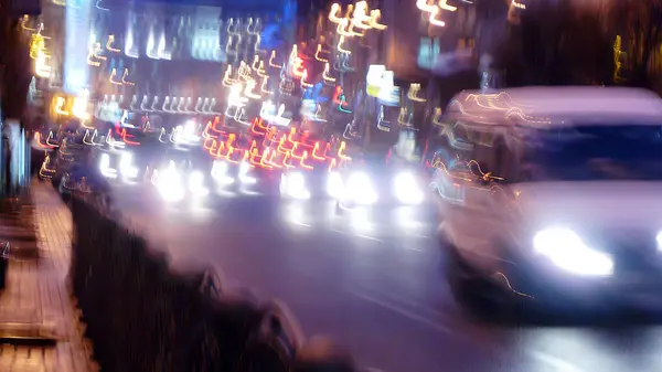 Bulanık tasvir gece boyunca canlı bir şehir ortamını gösteriyor. Beyaz bir araç hareketli caddede yürürken, araçların ve sokak lambalarının renkli ışıkları olay yerini aydınlatıyor. Genel etki bir hareket hissi yaratır.