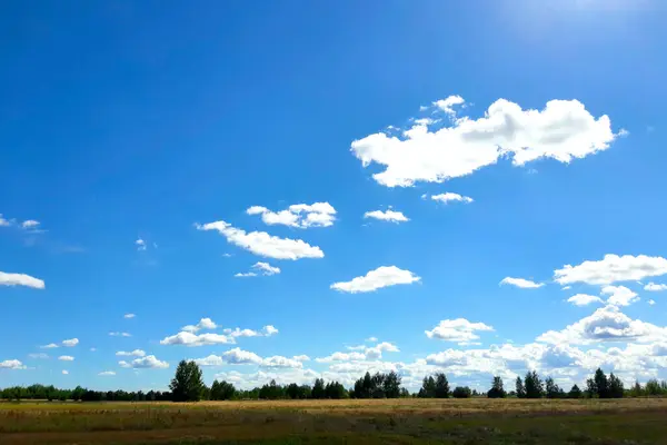 stock image A clear blue sky filled with fluffy white clouds stretches over a peaceful countryside landscape, illuminated by sunlight.