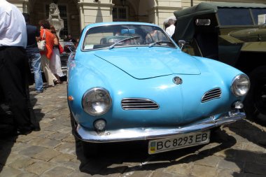 Lviv, Ukraine - June 28, 2014 A vintage blue car Volkswagen Karmann Ghia Coupe stands proudly in a charming square filled with spectators admiring various vehicles clipart