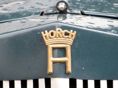 Lviv, Ukrayna - 14 Haziran 2015 Antik Horch ambleminin yakın çekimi yağmur damlalarıyla süslenmiş, klasik bir araba etkinliği sırasında yakalanan, otomotiv tarihi ve zanaatkarlığını kutlayan karmaşık metal işçiliğini ön plana çıkarıyor..