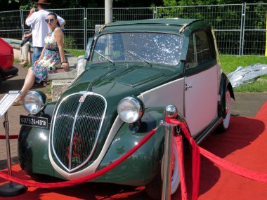 Lviv, Ukraine - June 5, 2016 A vintage two-tone car FIAT Topolino stands proudly red carpet at an outdoor event, while a woman in a floral dress admires its classic design under the warm summer sun. clipart