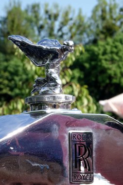 Lviv, Ukraine - June 6, 2017 A beautifully polished Rolls-Royce Phantom II hood ornament gleams in the sunlight during a car show. The background features greenery and event tents, creating a festive atmosphere. clipart