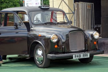 Lviv, Ukraine - June 2, 2018 A vintage black taxi Austin FX4 1989 models with one door open, parked at a lively event featuring classic cars under clear blue skies. clipart