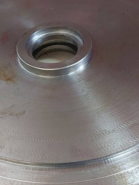 Paslanmaz çelik bir diskin yakın görüntüsü, pürüzsüz yüzeyi ve merkezi deliği vurguluyor, aletlerle dolu işlek bir metal atölyesinde çalışma tezgahında dinleniyor..