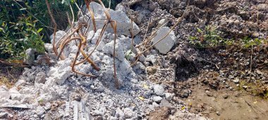 Beton moloz yığınları, inşaat sahasına saçılmış beton ve toprak, yakınlarda yeşil bitkiler yetişiyor ve yerde çamurlu toprak görünüyor..