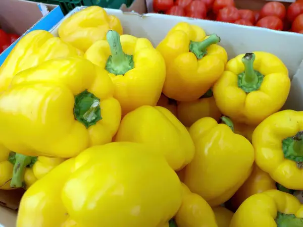 Parlak sarı tatlı biberler bir pazar kutusunda toplanır, canlı renklerini ve sağlıklı görünüşlerini diğer sebzeler arasında sergilerler..