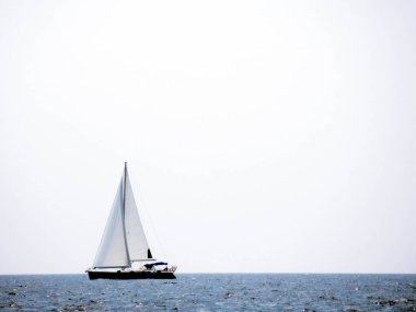 Yelkenleri açık bir yat güneşli bir günde sakin denizde yol alır. Suyun engin genişliği berrak bir gökyüzü altında uzanıyor ve sakin bir atmosfer yaratıyor..