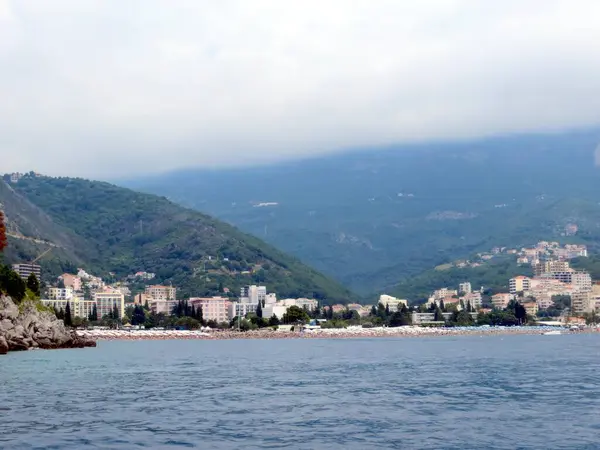 Budvas 'ın çarpıcı kıyı şeridinde oteller ve sakin bir marinanın yanında canlı bir sahil bölgesi, arka planda yemyeşil dağlar ve tepede bulutlar bulunur..