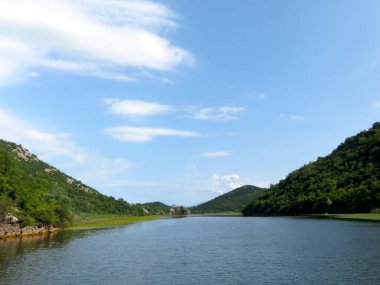 Berrak bir mavi gökyüzü altında, Skadar Gölü çevresindeki yemyeşil alanları yansıtır, gezginleri sakin sularını ve çarpıcı manzaralarını keşfetmeye davet eder..