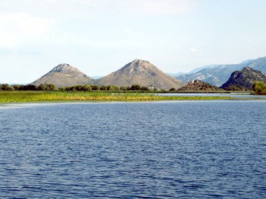Skadar Gölü Karadağ 'ın dingin güzelliğini yansıtıyor, berrak mavi bir gökyüzü altında sakin suların manzaralı dağlar ve yemyeşil alanlarla çevrili olduğu.
