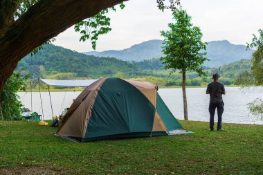 Gölde tatil yapan Asyalı erkek turist kampı, büyük ağaçların altındaki yeşil çimlerin üzerinde kamp kurma, brandaları yayma, güzel göl ve dağ manzarası, tatil aktiviteleri..