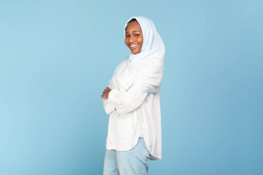 Başörtülü mutlu Afro-Amerikan kadın portresi. Mavi arka planda kollarını kavuşturmuş poz veriyor. Başörtüsü takmış, kameraya bakıp gülümseyen pozitif siyah kadın.