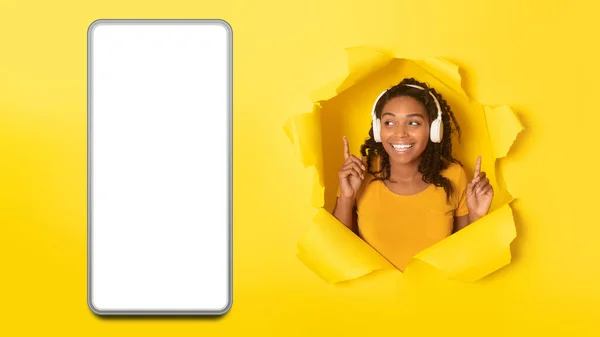 明るいアフリカ系アメリカ人女性が黄色のスタジオの背景にある黄色い穴を見て空の画面を持つ大きなスマートフォンの近くのヘッドフォンで音楽を聴いています パノラマ コラージュ — ストック写真