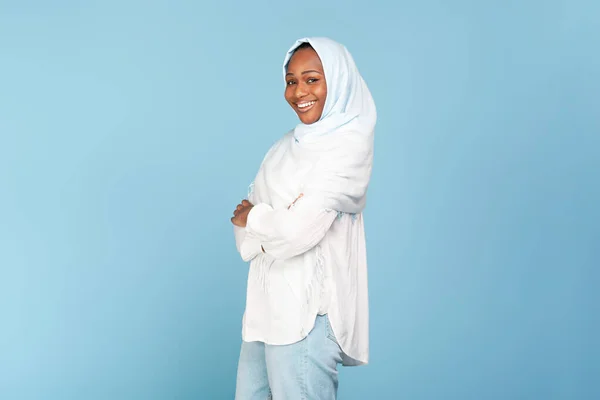 Portrait of happy african american lady in hijab posing with folded arms over blue background, positive black woman in headscarf looking and smiling at camera, free space