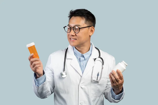乳白色のコートで中国の男性医師高齢者の笑顔 眼鏡薬の瓶を見て 青の背景に スタジオに孤立した 薬の選択 広告と提供 病気や医療の治療 — ストック写真