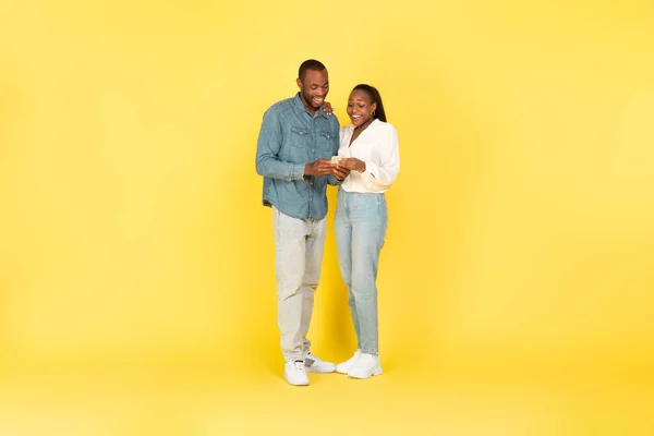 stock image Full Length Shot Of Black Couple Using Cellphone Texting Together Standing Over Yellow Background In Studio. Couple Using New Mobile Application. Technology And Gadgets