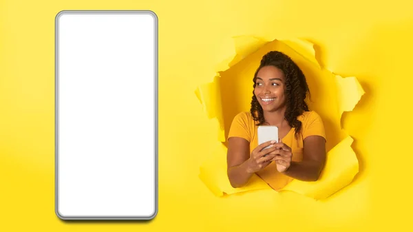 明るいアフリカ系アメリカ人女性が黄色のスタジオの背景に空白の画面を持つ大規模なスマートフォンを脇に見てトーンホールで携帯電話のポーズを使用して 新しいモバイルアプリケーション モックアップパノラマ — ストック写真