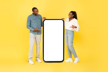 Heyecanlı Siyah Eşler, Geniş Cep Telefonu Ekran Reklamında Büyük Cep Telefonu Reklamını İşaret Ediyor Sarı Stüdyo Arkaplanının Üzerinde Birlikte Poz Veriyorlar. Model, Tam Uzunluk