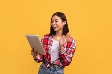 Heyecanlı Asyalı kadın videosu, dijital tabletle web kamerasına el sallayarak sarı stüdyo arkaplanı üzerinden çevrimiçi iletişim kuruyor. Modern iletişim konsepti