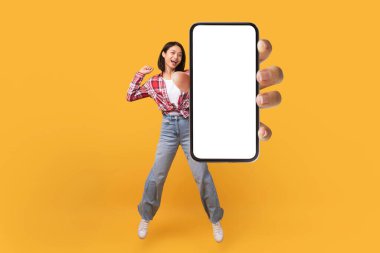 Mutlu Koreli kadın sarı stüdyo arka planında sıçrarken boş akıllı telefon ekranı gösteriyor, kolaj, maket. Havalı mobil uygulama ya da web sitesi öneren heyecanlı bir kadın