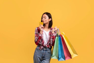 Alışveriş zamanı. Genç Asyalı bayan müşteri ellerinde renkli alışveriş çantaları ve öpüşme bankası kredi kartı tutarken sarı stüdyo arka planında izole bir şekilde duruyor.